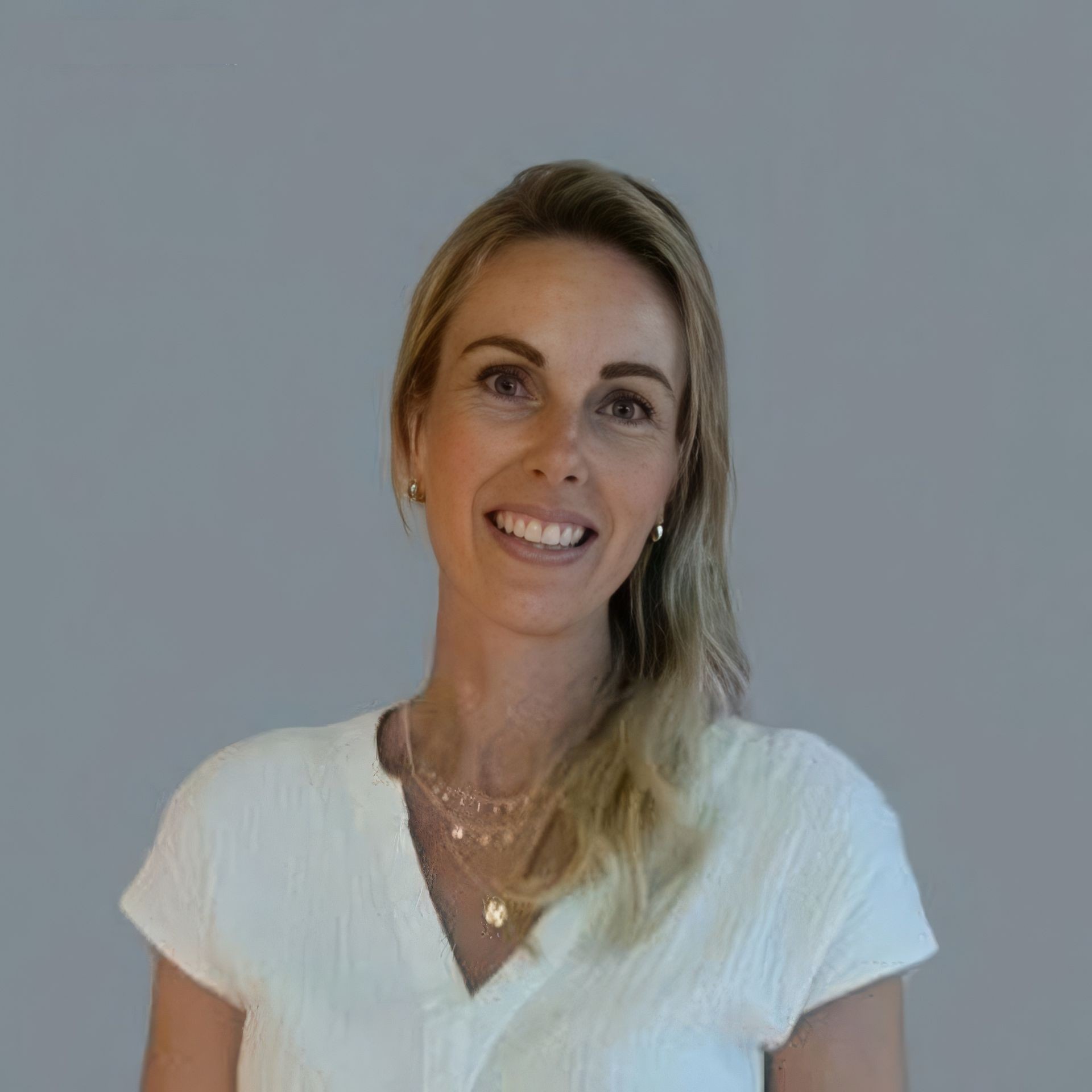 A woman with blonde hair wearing a white top and layered necklaces against a plain background.